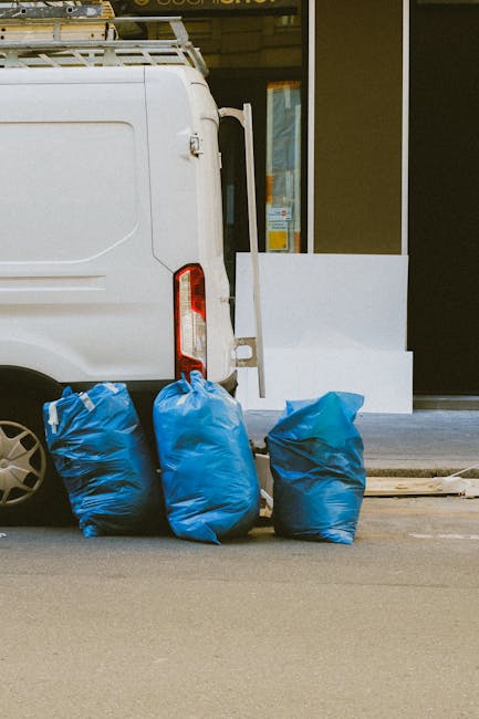 Two dark green, wheelie bins with closed lids are situated on a paved sidewalk at the edge of a street, close to a curb. The bins are made of plastic, with a textured finish, and are positioned side b